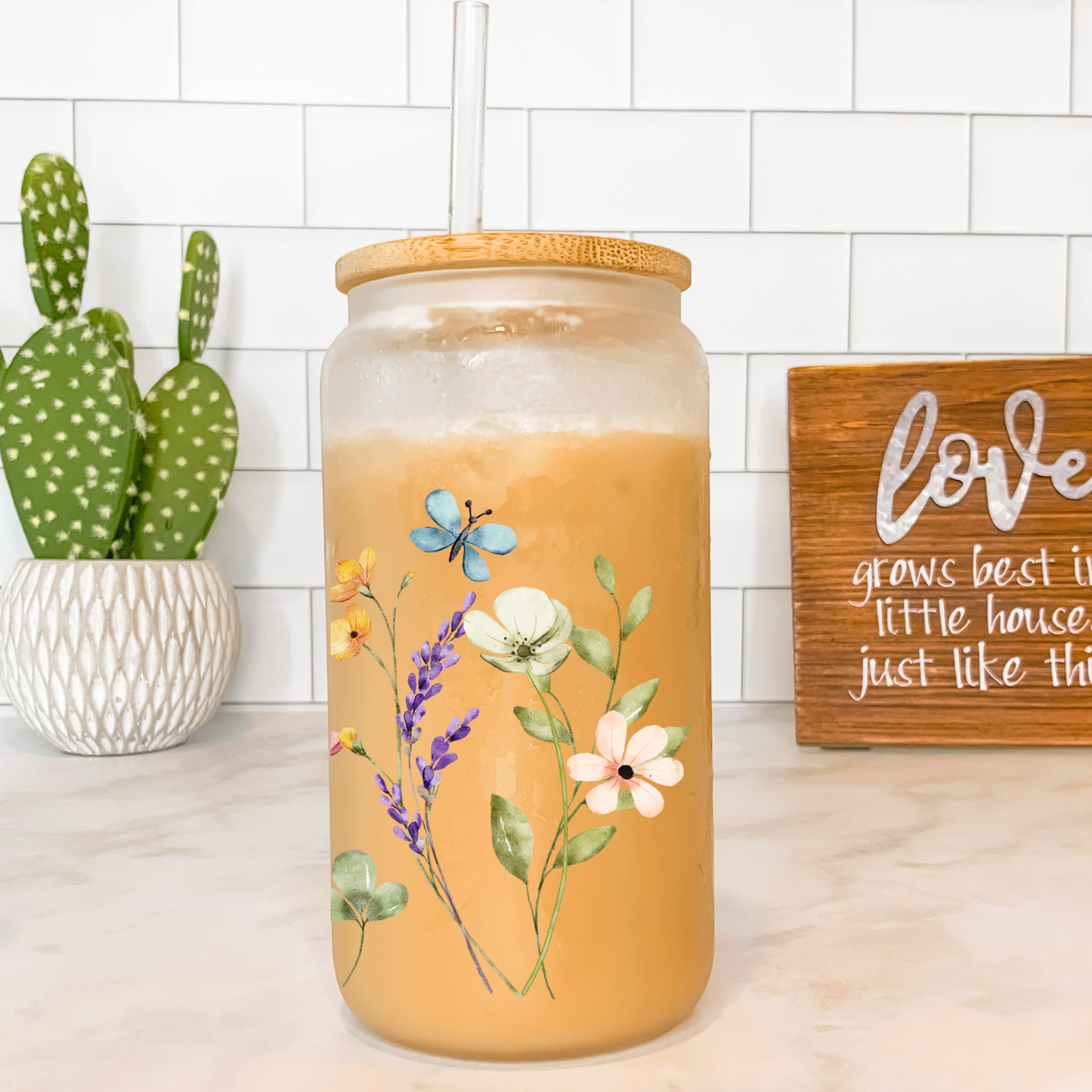 Decorative jar with floral design on a countertop with a cactus plant and 'love' sign in the background.