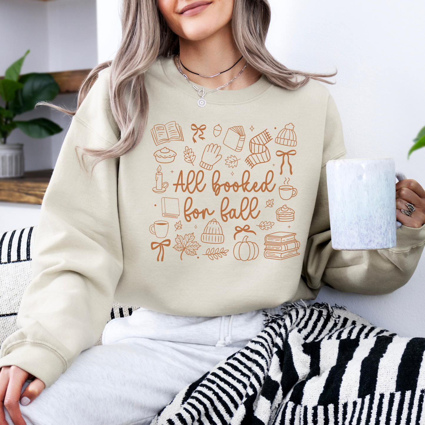 Person wearing a beige sweatshirt with 'All booked for fall' text and autumn illustrations, holding a mug.