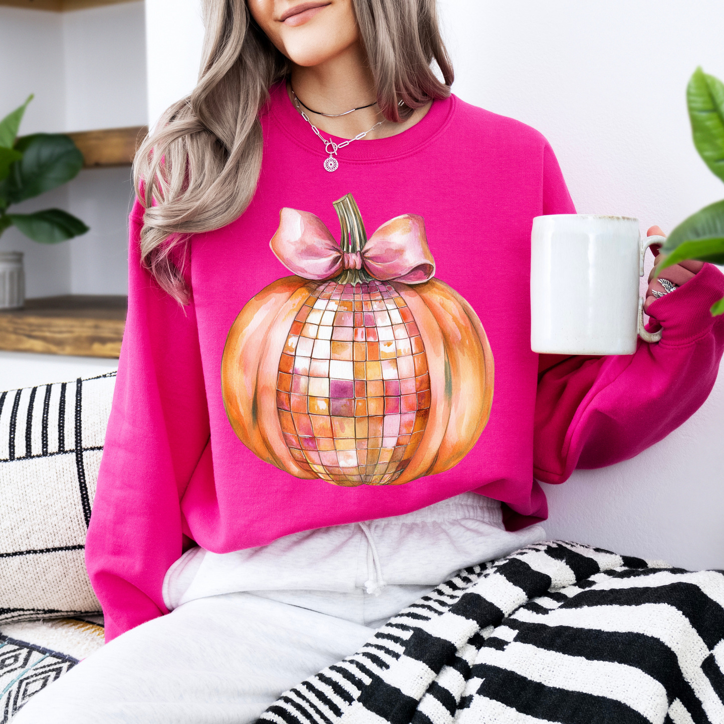 Person wearing a pink sweatshirt with a disco pumpkin design, holding a white mug.