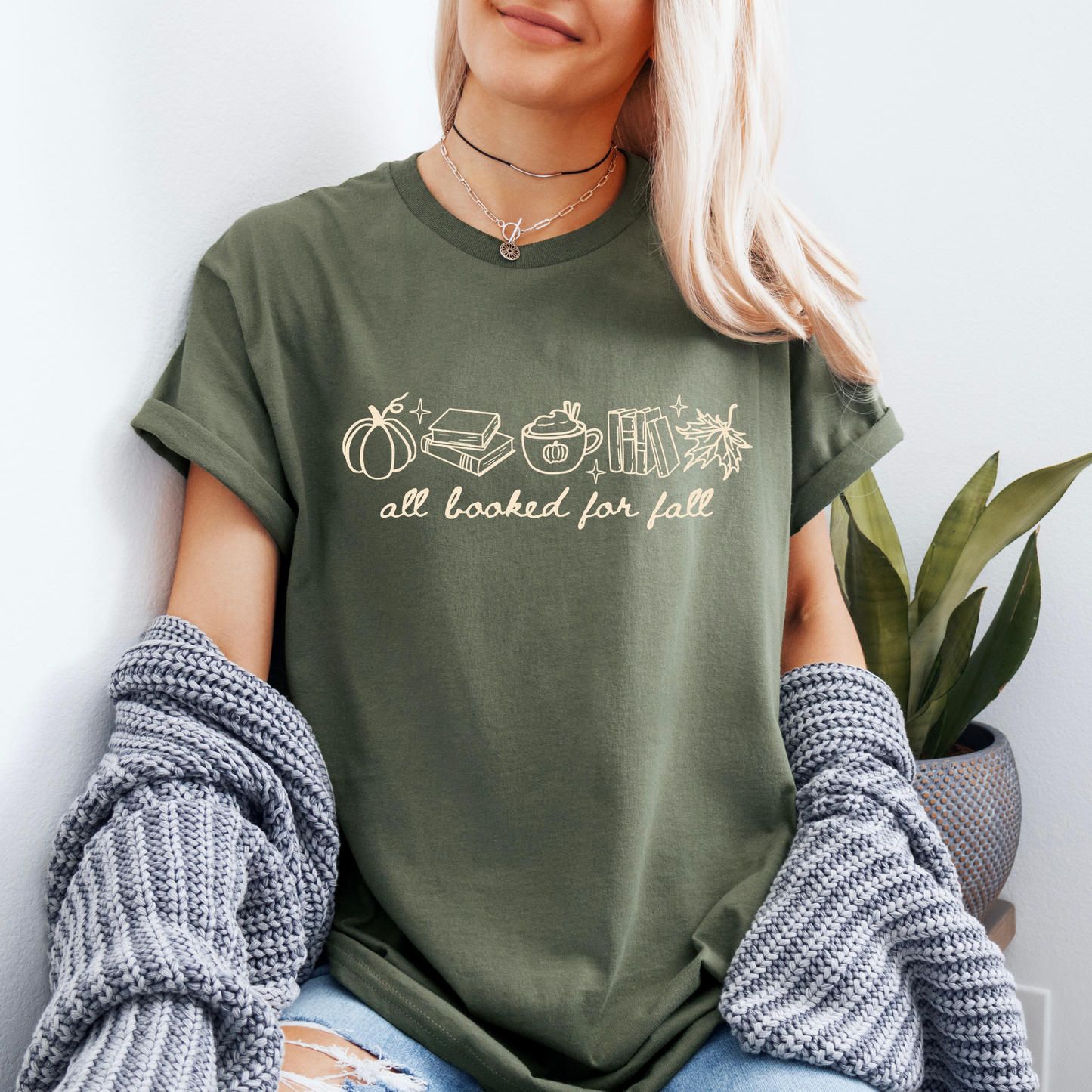 A woman wearing an olive green graphic t-shirt with a fall-themed illustration and the text 'all looked for fall'. She has a gray knit sweater off her shoulders and is leaning against a white wall next to a potted plant.