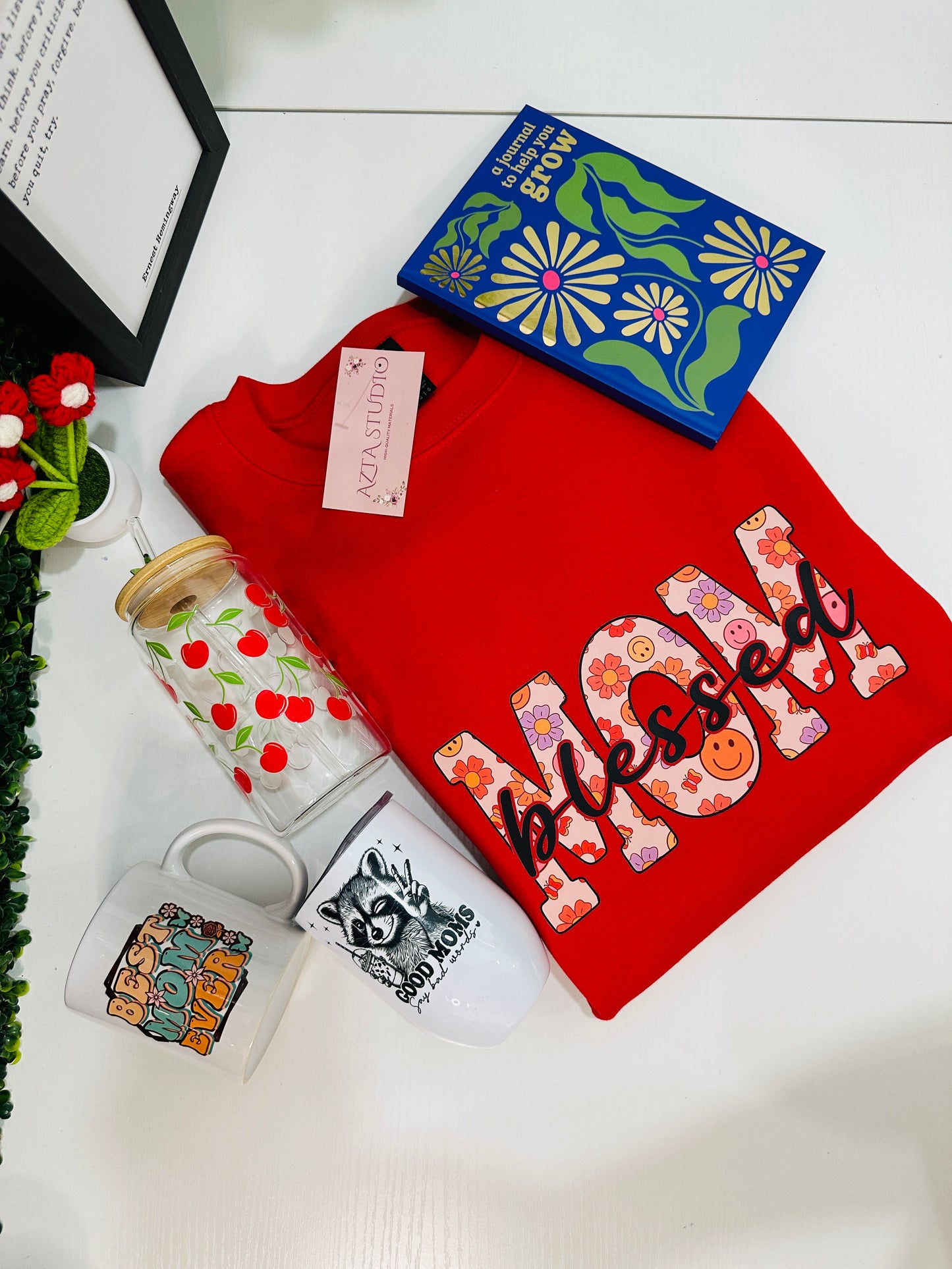 Red sweatshirt with 'Messed Up Mom' design, mug, tumbler, and decorative box on a white surface.