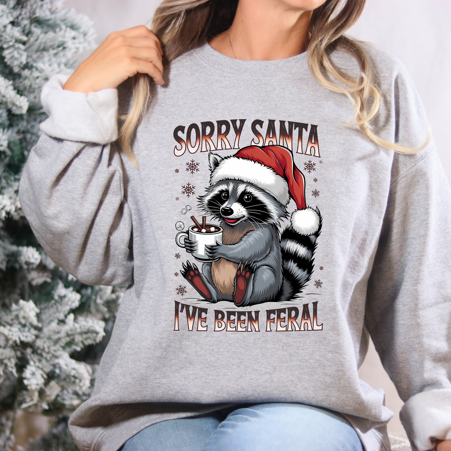 Person wearing a gray sweatshirt with a raccoon graphic and text in front of a Christmas tree.