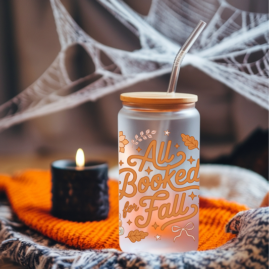 A frosted glass can-shaped tumbler with a bamboo lid and metal straw, printed with the phrase 'All Booked for Fall' in orange script with autumn leaves and stars. The tumbler is styled on an orange knit blanket next to a lit black candle, with a blurred background of spiderwebs and warm lighting.