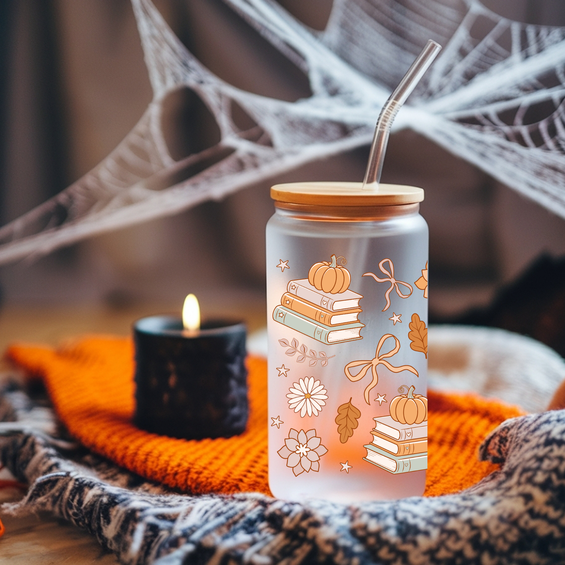 A frosted glass tumbler with a bamboo lid and metal straw, decorated with illustrations of stacked books, pumpkins, bows, and autumn leaves. It is styled on an orange knit blanket next to a lit candle, with cobweb decorations in the background.