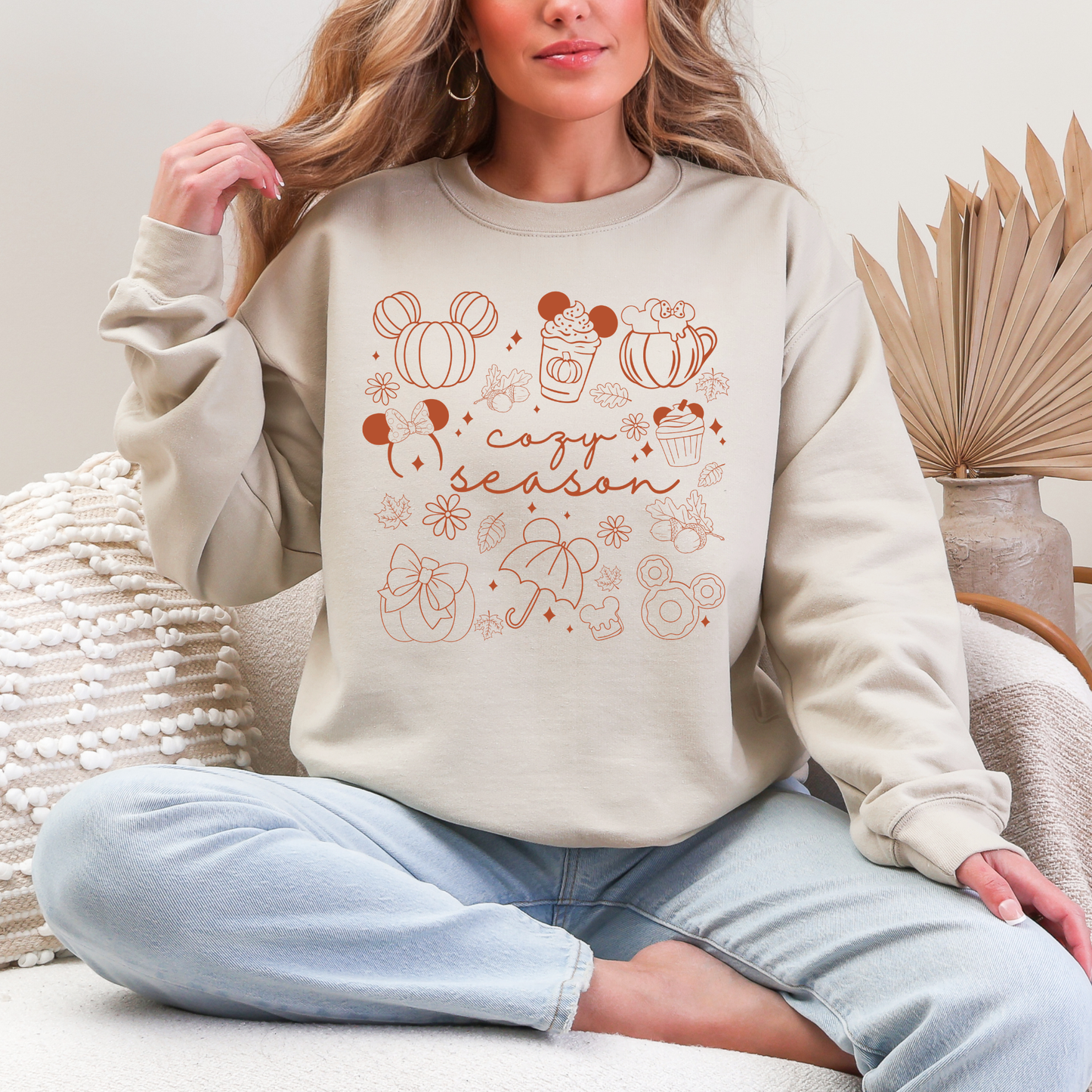 A woman wearing a beige crewneck sweatshirt with a 'cozy season' graphic featuring pumpkin, cupcake, and Mickey Mouse designs, paired with light blue jeans in a cozy indoor setting.