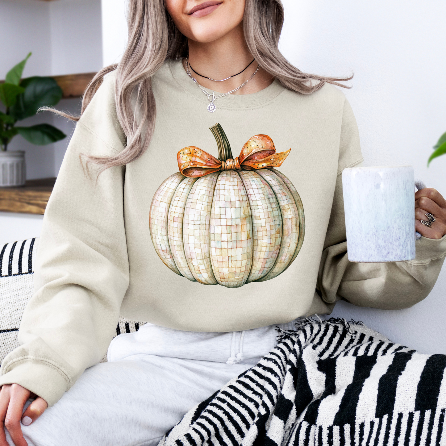 Person wearing a beige sweatshirt with a pumpkin design, holding a white mug.