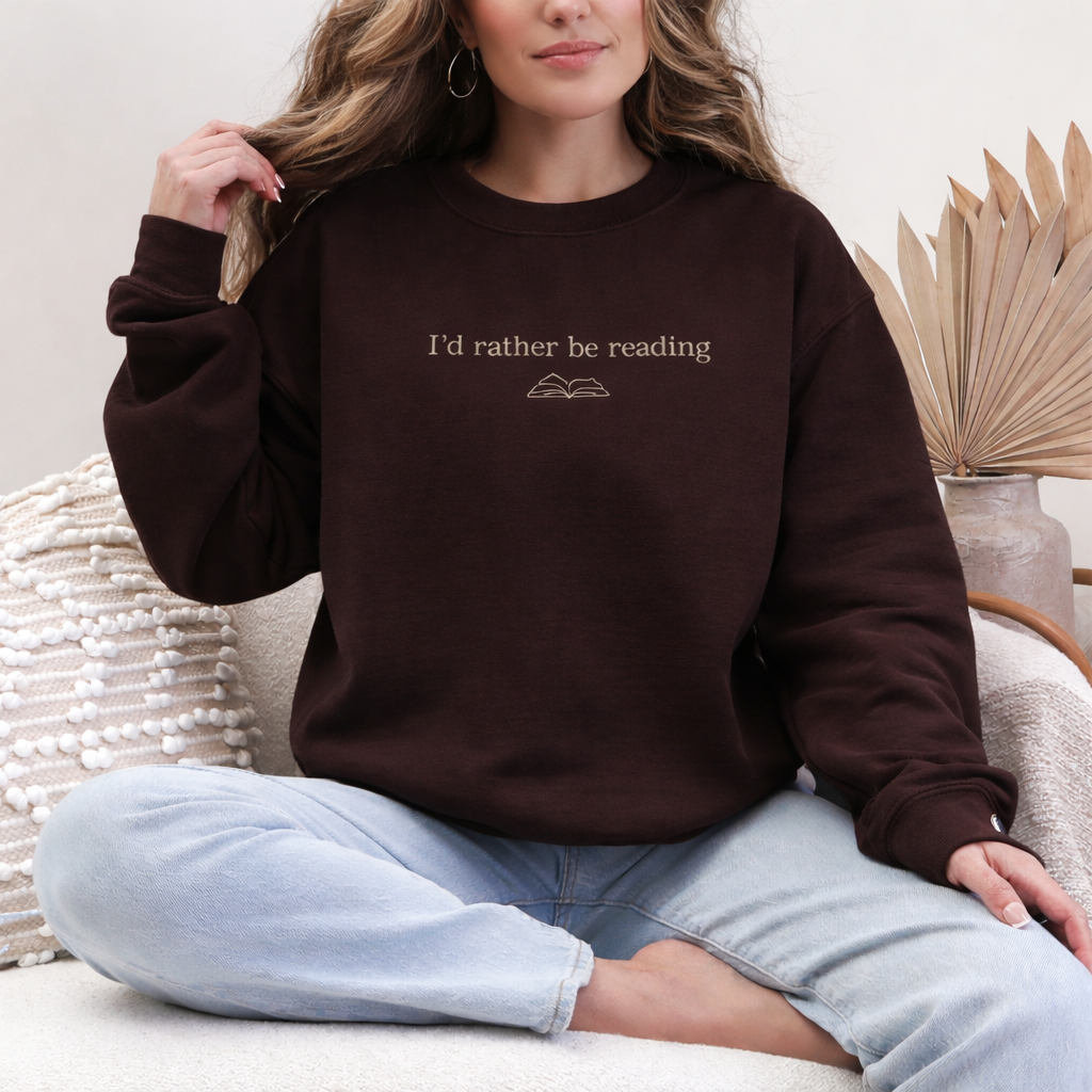 Person wearing a brown sweatshirt with 'I'd rather be reading' text, sitting on a white surface.