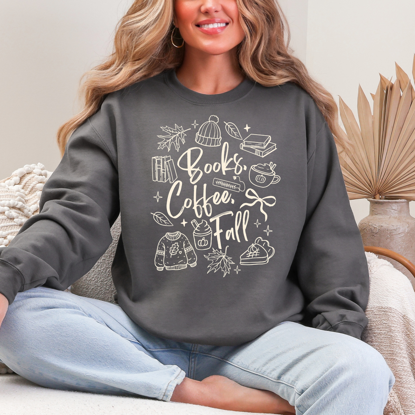 A woman sitting cross-legged on a couch wearing a dark gray crewneck sweatshirt with a white graphic that reads 'Books, Coffee, Fall' surrounded by autumn-themed illustrations. She is wearing light blue jeans in a cozy indoor setting.