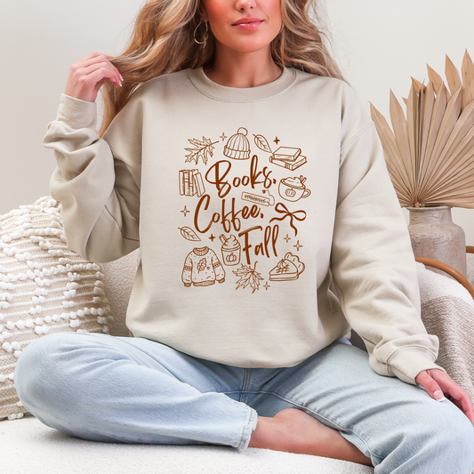 A woman sitting cross-legged on a couch wearing a beige crewneck sweatshirt with a brown graphic that reads 'Books Coffee Fall' surrounded by autumn-themed illustrations. She is wearing light blue sweatpants in a cozy indoor setting.