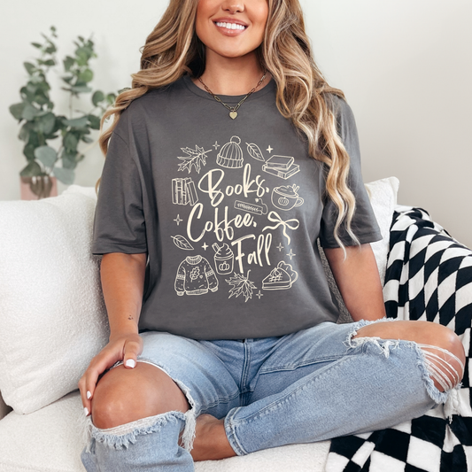 A woman sitting on a white couch wearing a dark gray graphic t-shirt with 'Books, Coffee, Fall' written in white script surrounded by autumn-themed illustrations, paired with distressed light blue jeans.