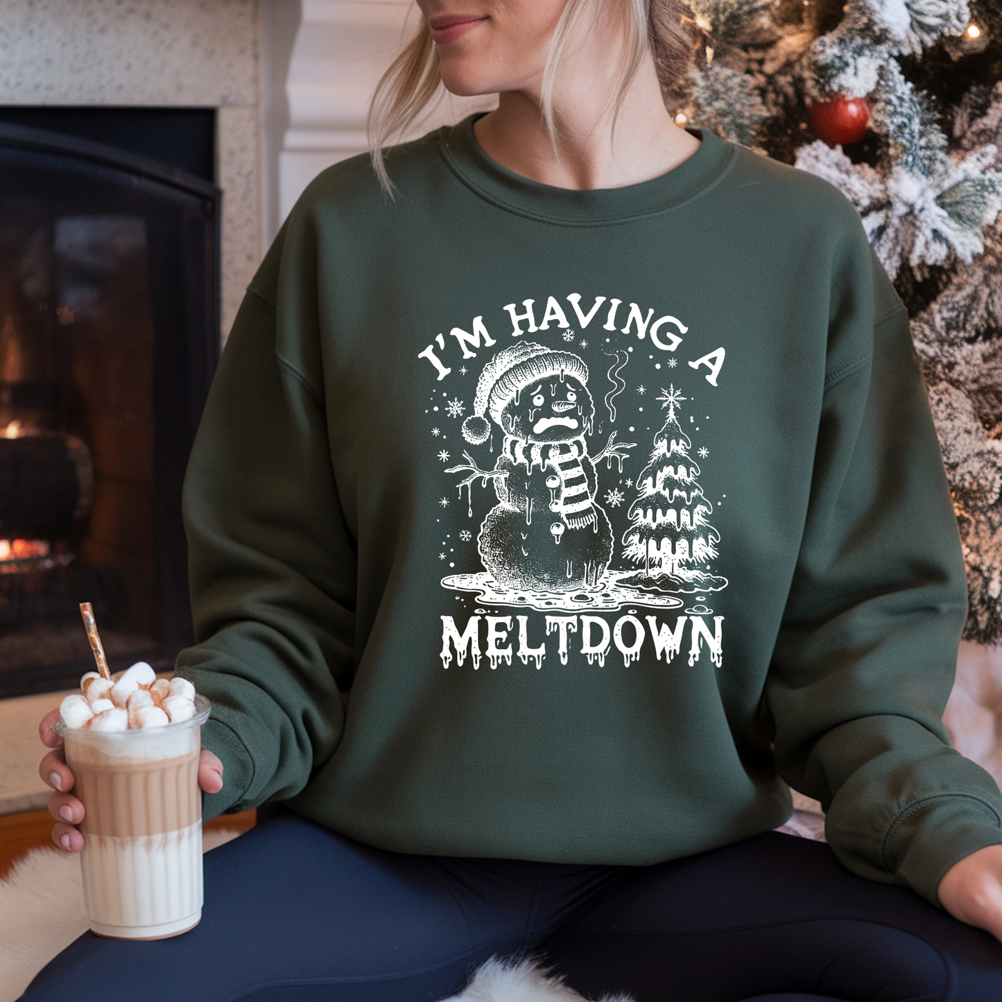 Person wearing a green sweatshirt with a snowman graphic and text, holding a hot chocolate drink.