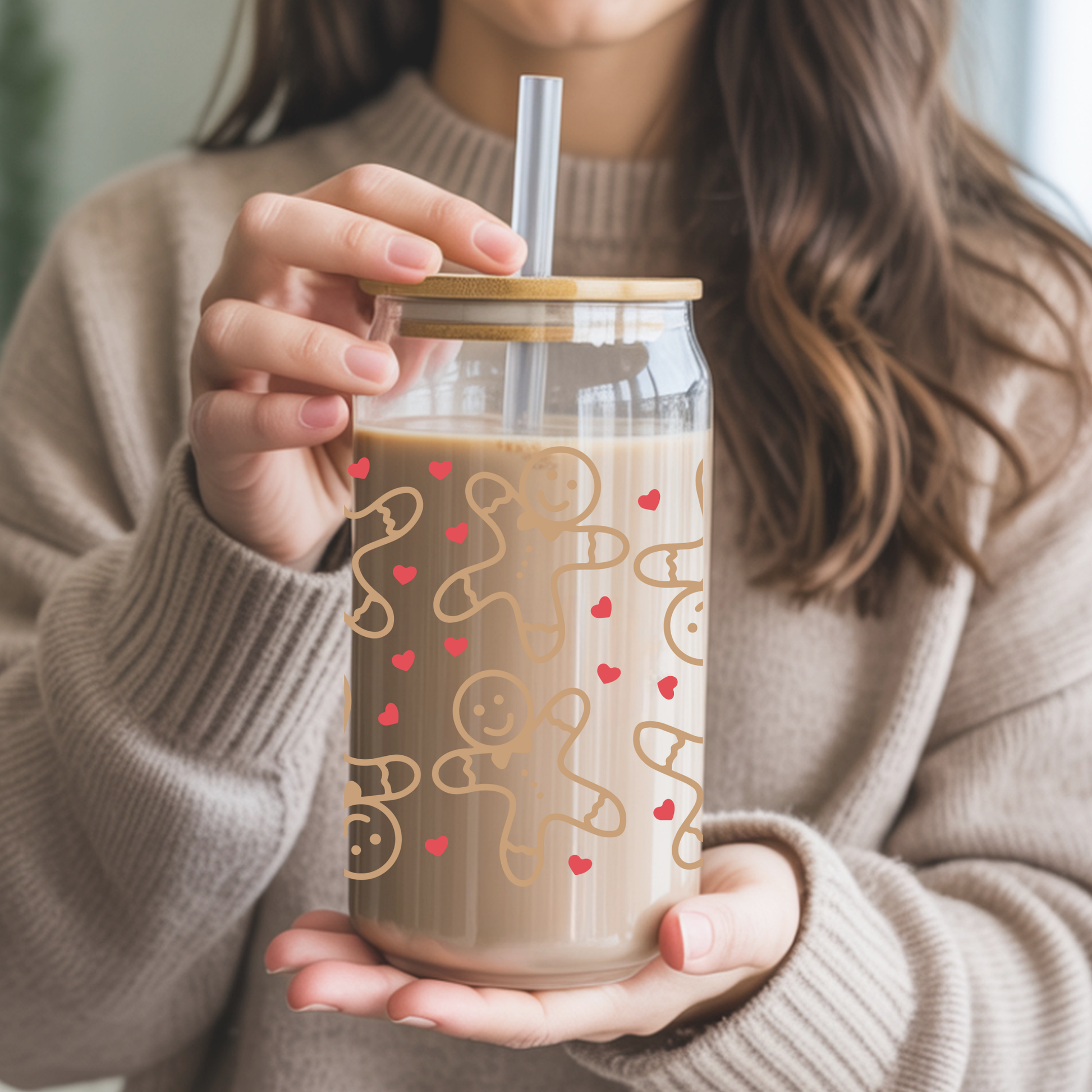 Person holding a glass mug with a lid and straw, featuring gingerbread men and heart designs.