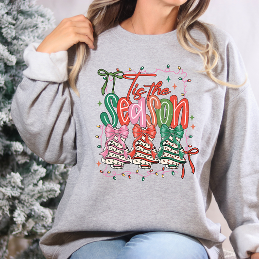 Person wearing a gray sweatshirt with 'Tis the Season' design in front of a Christmas tree.