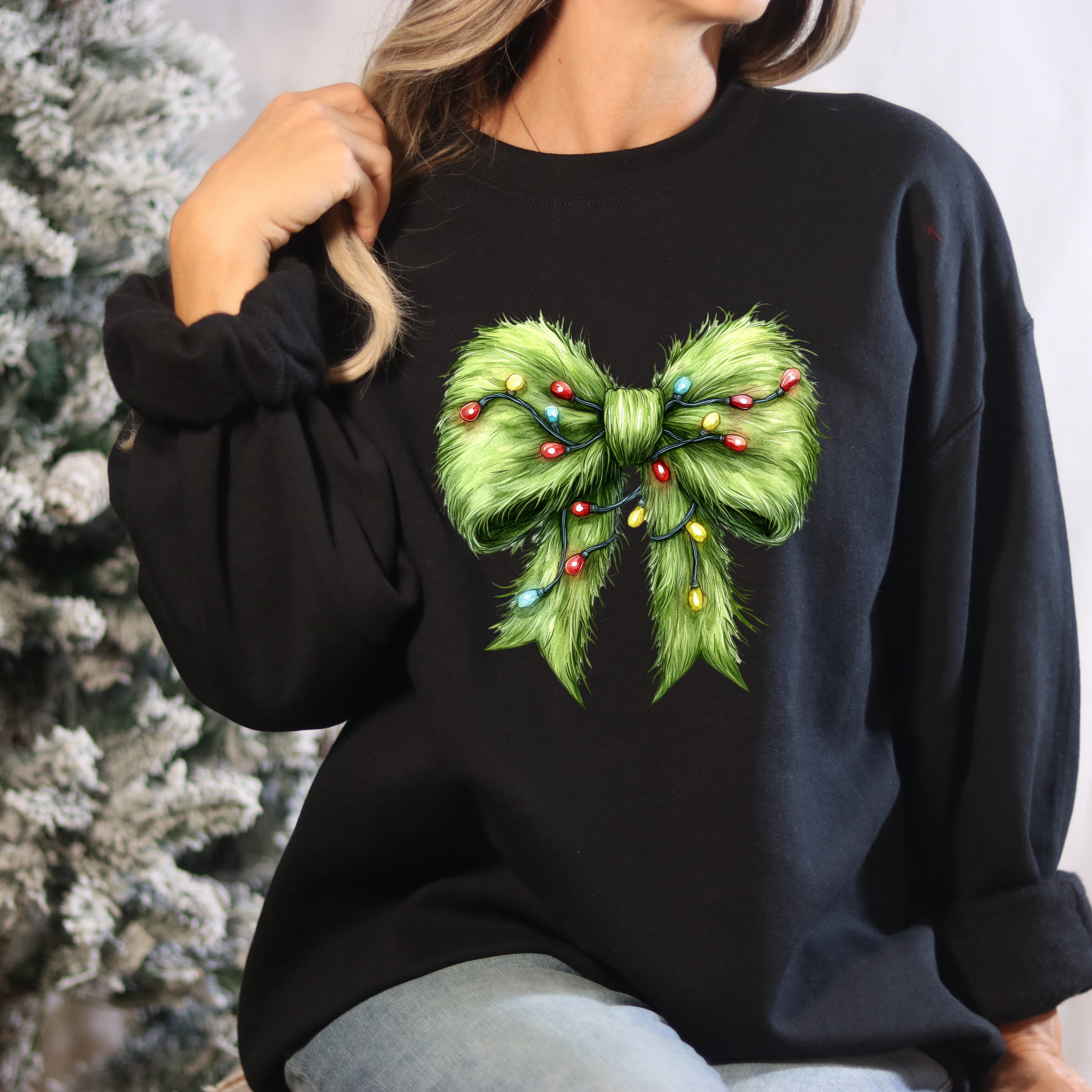 Black sweatshirt with a green bow design featuring colorful lights, worn by a person against a blurred Christmas tree background.