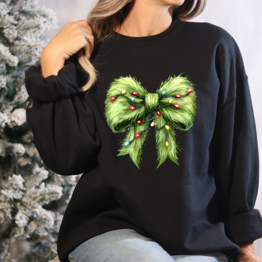 Black sweatshirt with a green bow design featuring colorful lights, worn by a person against a blurred Christmas tree background.