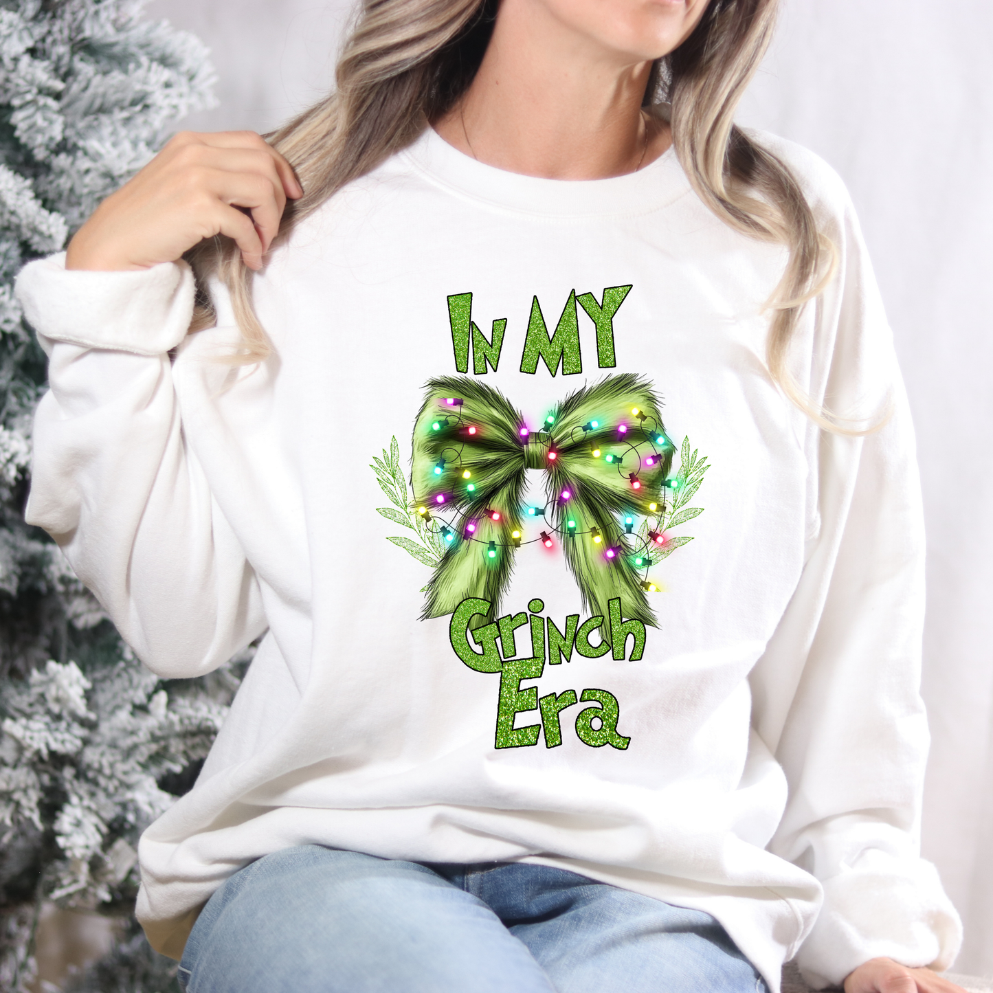 Person wearing a white sweatshirt with a festive design and text, sitting in front of a Christmas tree.