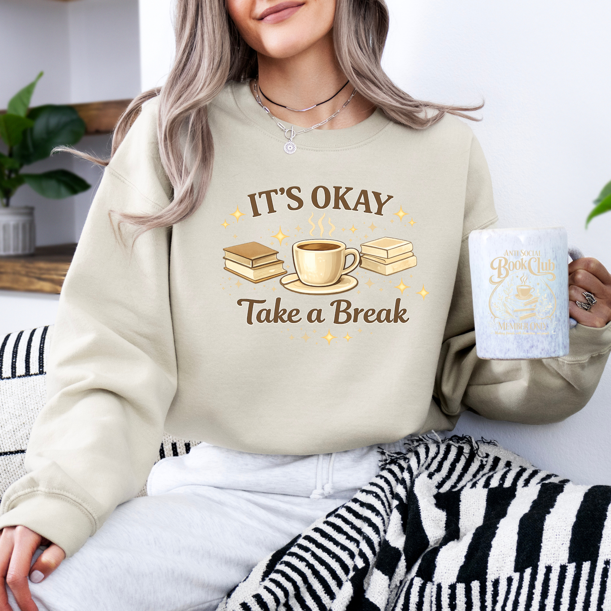 Person wearing a beige sweatshirt with 'It's Okay, Take a Break' text and holding a mug.