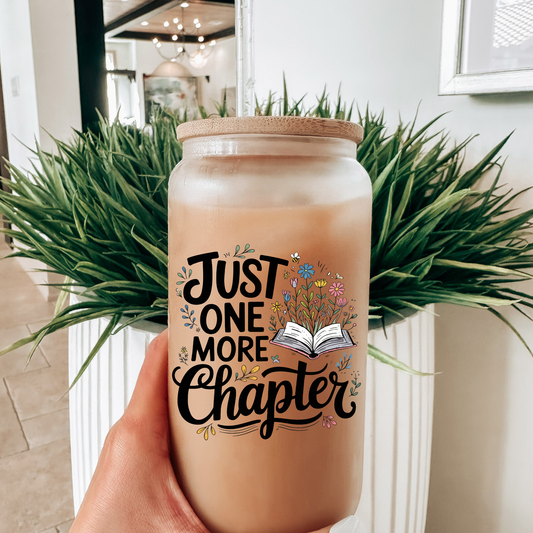 Hand holding a glass mug with 'Just One More Chapter' text and floral design, against a blurred indoor background.