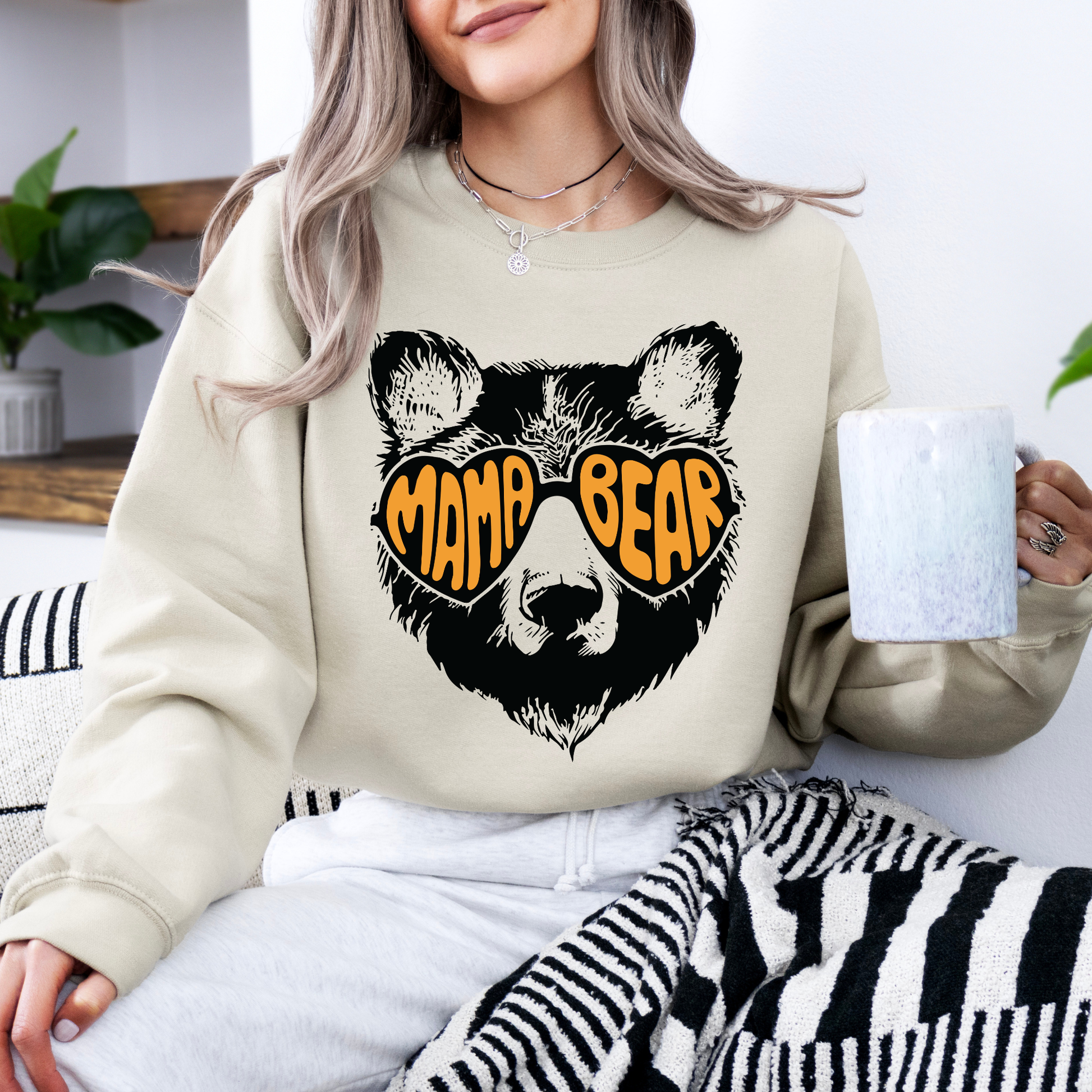 Person wearing a beige sweatshirt with a bear graphic and 'Mama Bear' text, holding a mug.