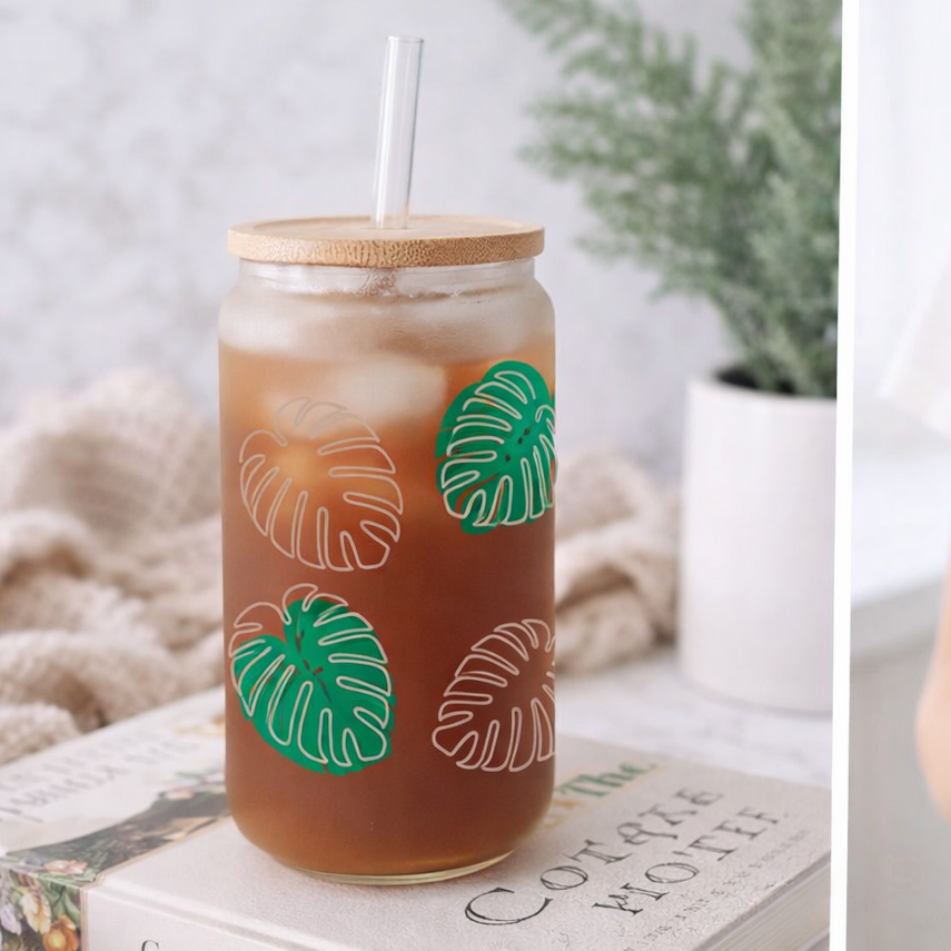 Mason jar with iced drink and leaf design held by a person, next to a book on a surface.