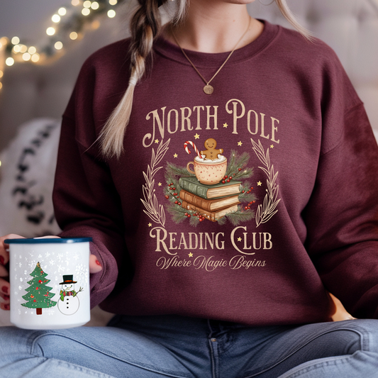 Person wearing a maroon sweatshirt with 'North Pole Reading Club' design, holding a mug.