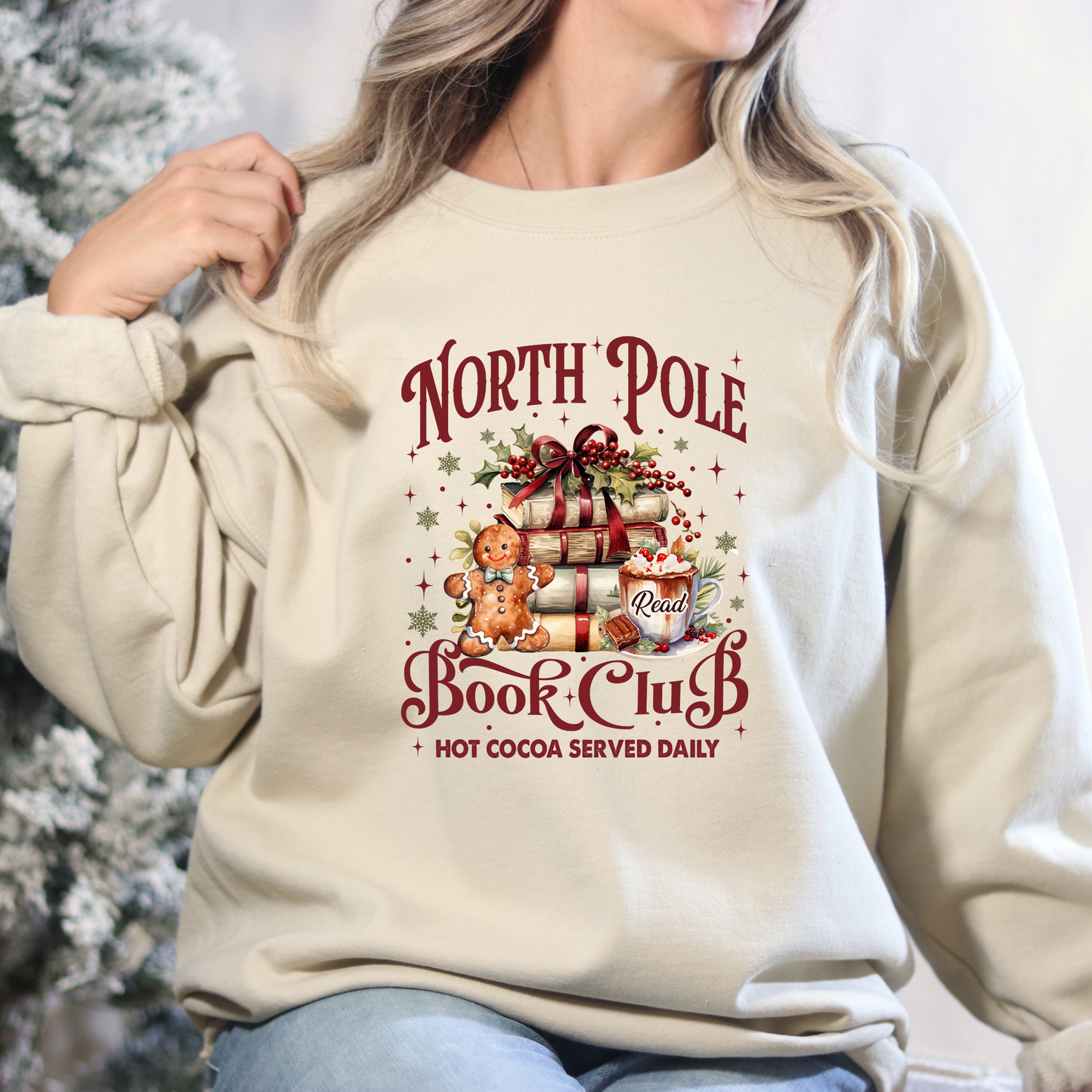 Person wearing a beige sweatshirt with 'North Pole Book Club' design, Christmas tree in the background