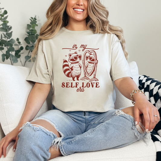 Woman wearing a t-shirt with a raccoon design and 'self love' text, sitting on a couch.