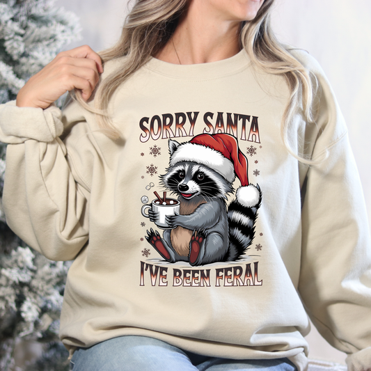 Beige sweatshirt with a raccoon wearing a Santa hat and text 'Sorry Santa I've Been Feral' against a blurred snowy background.
