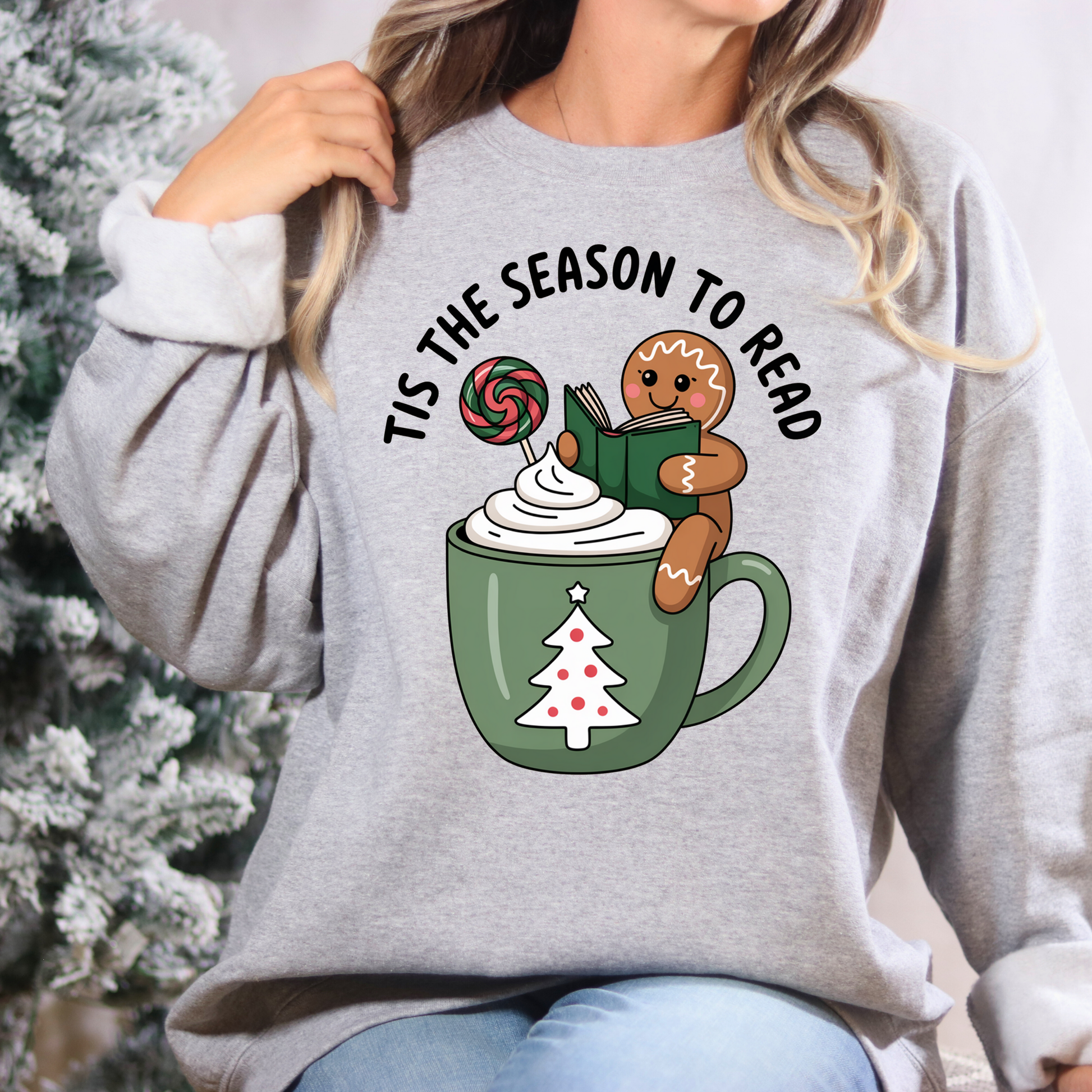 Gray sweatshirt with Christmas-themed design featuring a mug, gingerbread man, and text 'Tis the Season to Read' against a blurred Christmas tree background.