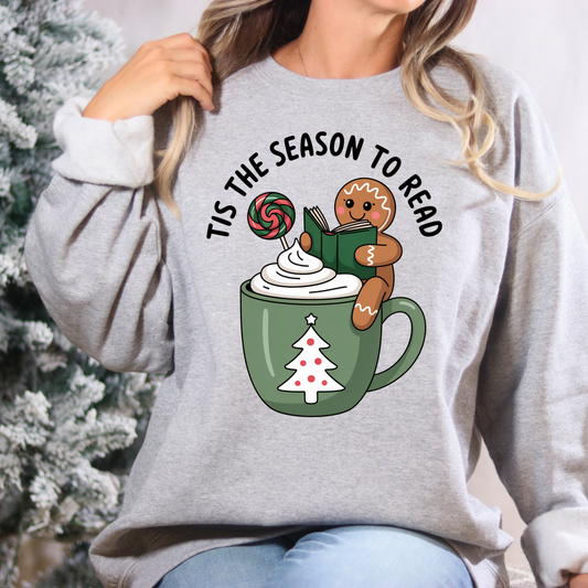 Gray sweatshirt with Christmas-themed design featuring a mug, gingerbread man, and text 'Tis the Season to Read' against a blurred Christmas tree background.